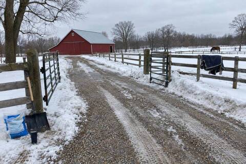 A plowed gravel lane leading to a red barn, a fenced paddock gate cleared on both sides, a horse blanket on the fence and snow-covered pasture beyond