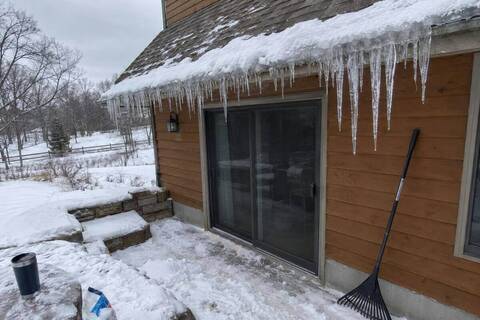 A ridge of ice and icicles along a shingled eave above a walkout basement patio door, snow on the roof above and on the steps below, a roof rake against the wall