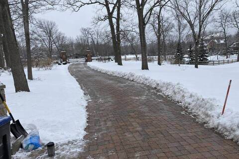 A long brick-paver driveway plowed in a single lane, curving through bare oaks toward an iron entrance gate, deep snow on both shoulders