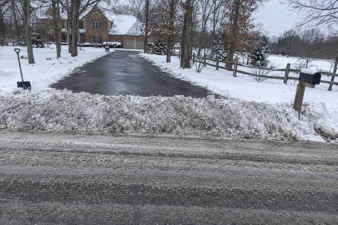 A dense ridge of plowed snow left across the foot of an asphalt driveway where it meets a two-lane road with no curb, plow tracks in the road and a mailbox post at the edge