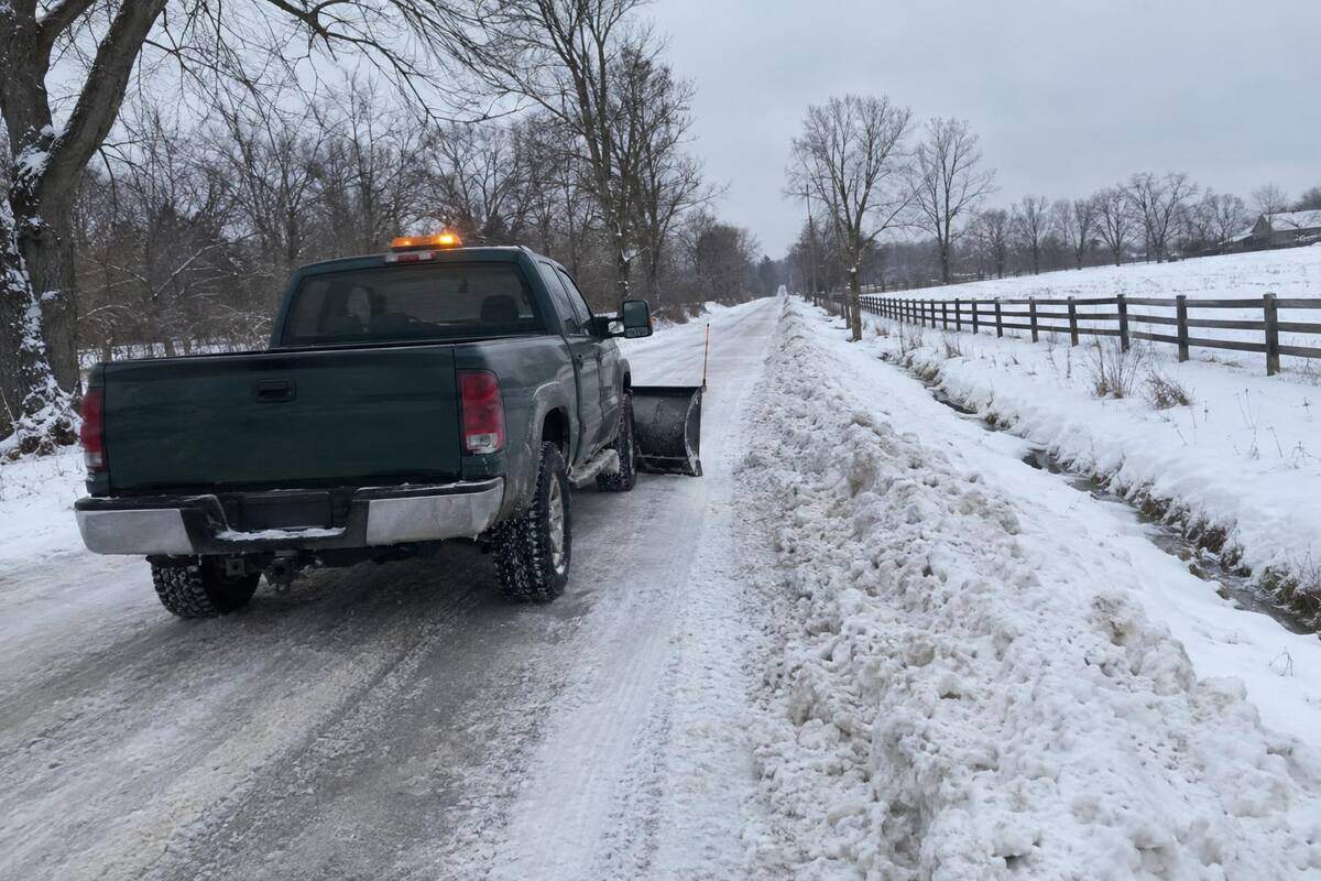 A dark green pickup with a straight plow blade seen from behind on a snow-covered rural road with no curb, amber light on the cab, the road ahead plowed one lane wide between bare trees