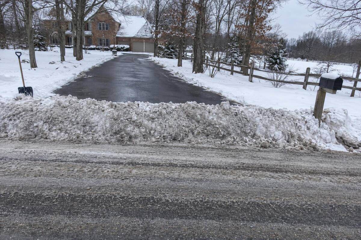 A dense ridge of plowed snow left across the foot of an asphalt driveway where it meets a two-lane road with no curb, plow tracks in the road and a mailbox post at the edge